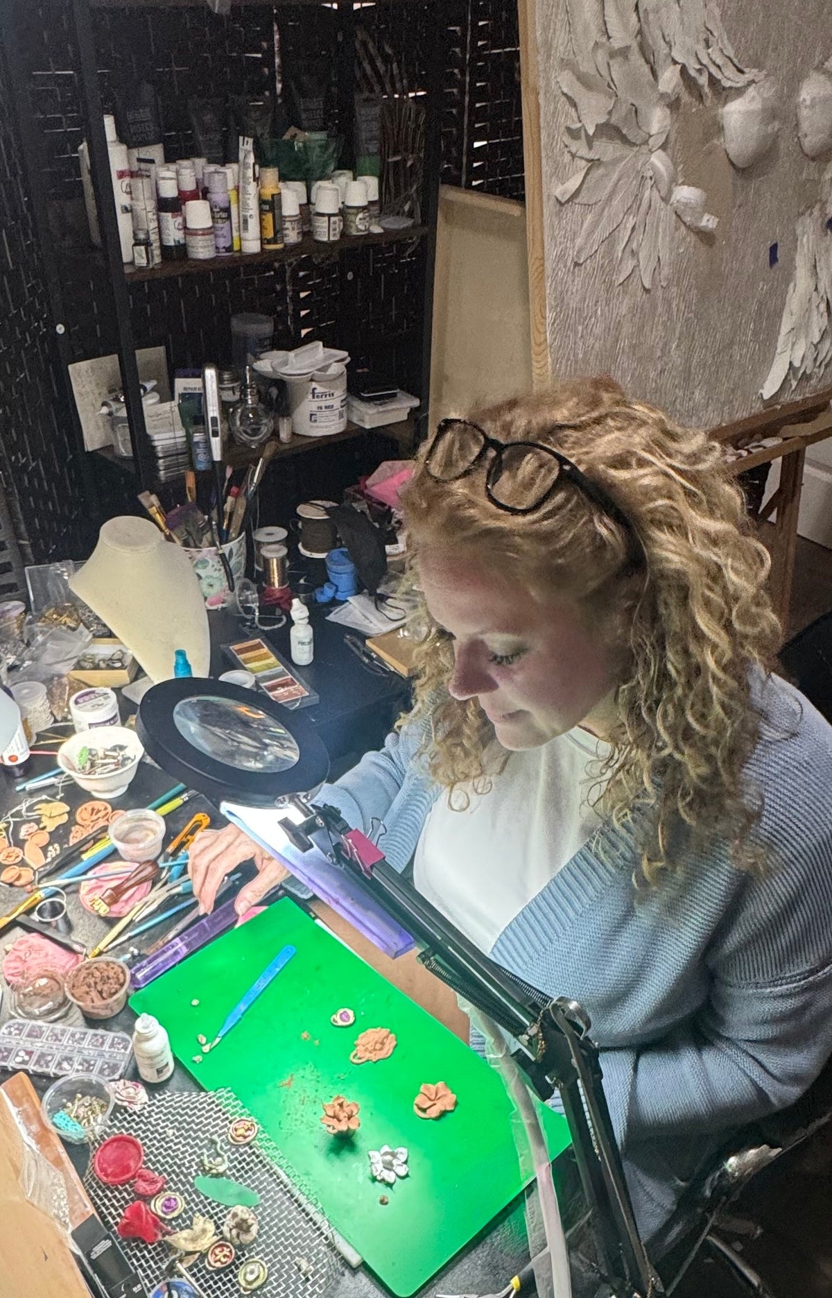 A woman with curly blonde hair and glasses sits at a cluttered craft desk, working under a bright lamp. The desk is covered in various small craft supplies, beads, and tools, and the background shows shelves filled with paintbrushes and art materials.